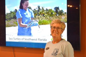 A woman with short blonde hair and glasses stands in front of a sea turtle presentation